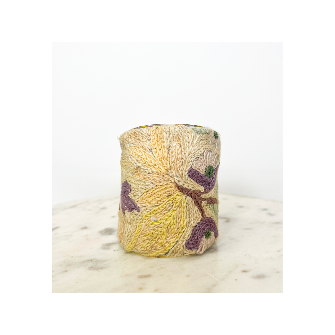 A vintage brass cuff bracelet with an embroidered design featuring yellow and purple patterns, set on a light-colored surface against a white background.
