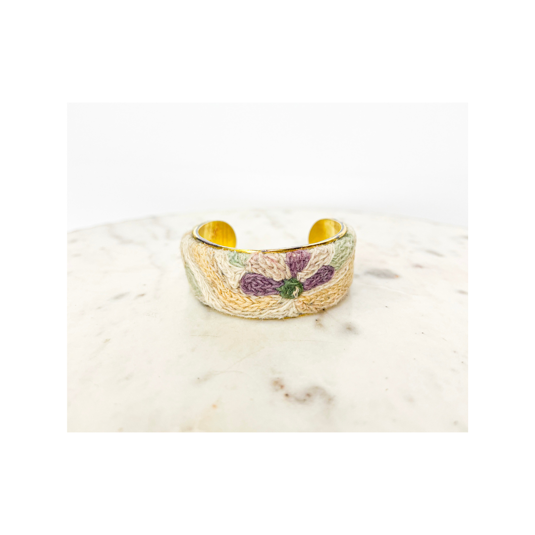A gold-colored, wide open vintage brass cuff bracelet with floral embroidery, placed on a white marble surface.