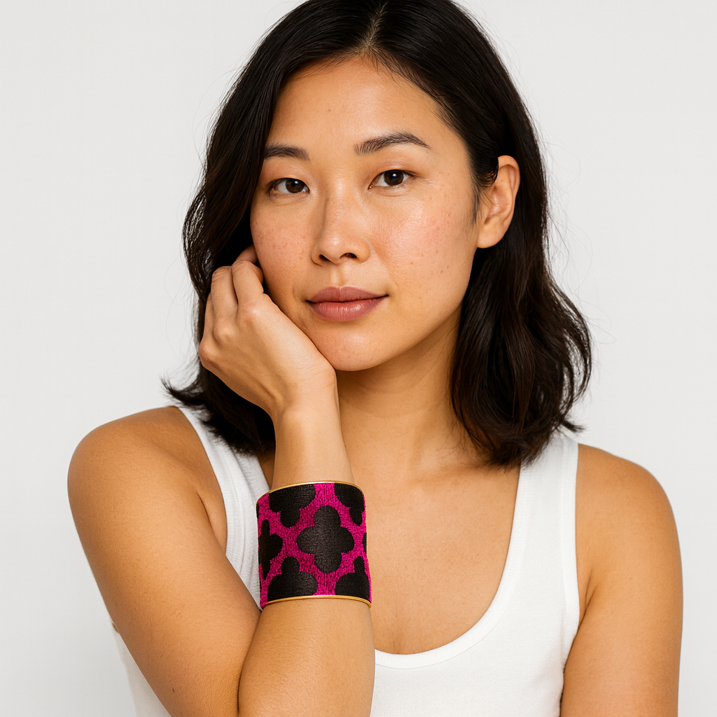 Close-up of a woman’s wrist and lower face, wearing the Redd Dott “Syntche” 3″ wide fabric cuff. The bracelet features a vivid fuchsia ground with oversized matte-black quatrefoil (clover) motifs, shown with slim matte-brass edges and an open-back frame as her hand rests at her cheek.