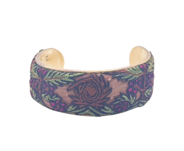 The Dawn bracelet by Redd Dott Jewelry showcases intricate floral embroidery in green and burgundy tones, stitched onto a vintage brass base. A handcrafted design blending elegance with rustic charm.