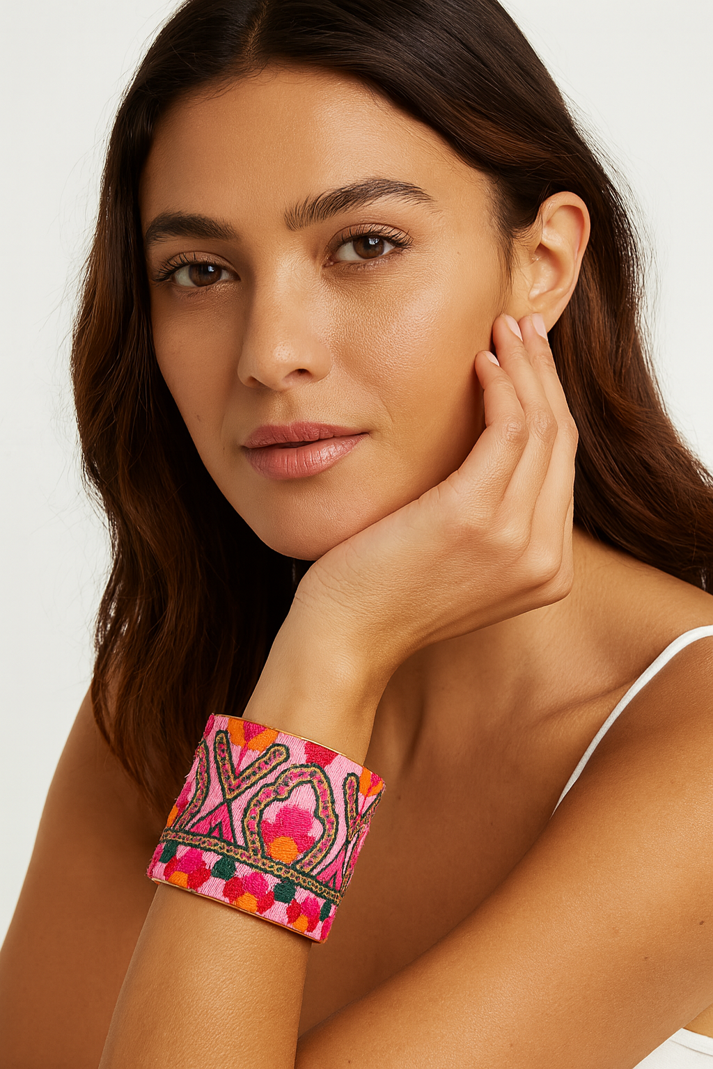 Close-up of a woman’s wrist and lower face, wearing the Redd Dott “Fiona” 2″ wide fabric cuff. The vibrant bubblegum-pink cotton is embroidered with looping emerald-green “XO” motifs and sprinkled with coral-orange stitch-dots, all framed by a slender open-back brass shell as her hand supports her cheek.