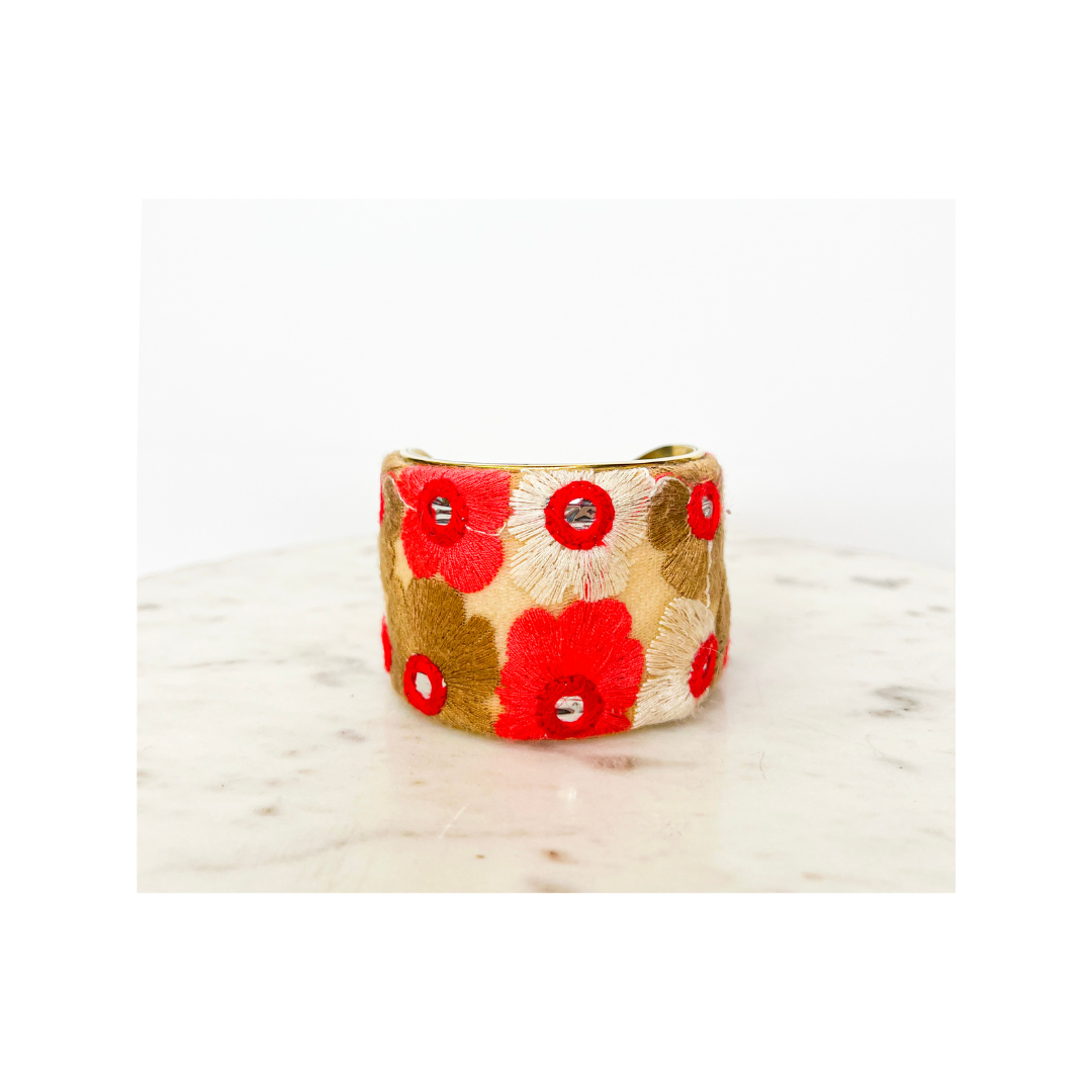 An embroidered bracelet with a vintage brass base, intricately adorned with handwoven flowers of red, cream and gold with mirrored center accents sits on top of a marble table.