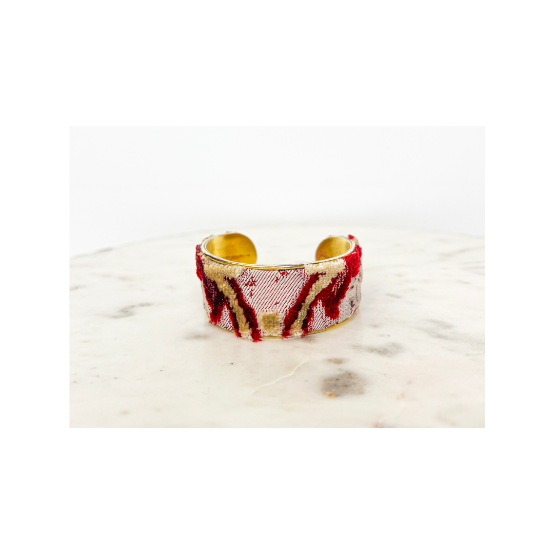 A gold cuff bracelet with a wide band and a textured, pink and red marbled design on its exterior, resembling the intricate patterns of a floral cuff bracelet, placed on a white marble surface.