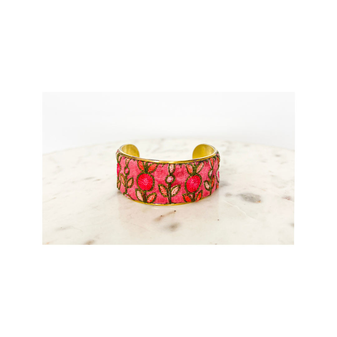 A pink floral cuff bracelet with gold accents and floral embroidery detail, displayed on a white marble surface.