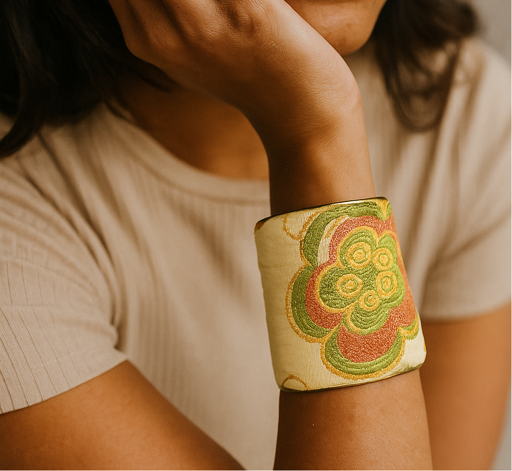 A female model’s wrist adorned with a 3-inch raw brass cuff bracelet featuring ivory fabric embroidered in coral and green floral patterns—an eye-catching one of a kind jewelry statement among cuff bracelets for women.