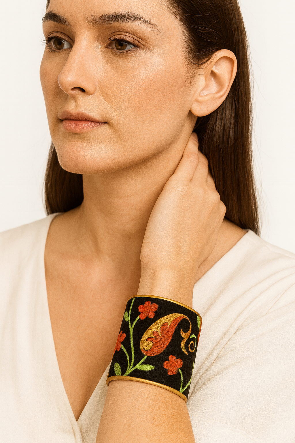 Close-up of a woman’s wrist and lower face, wearing the Redd Dott “Penelope” 3″ wide fabric cuff. The deep black cotton base is richly embroidered with oversized golden-yellow and burnt-orange paisley leaves, interspersed with tiny chartreuse-green stems and petite orange blossoms, all framed by a gently curved open-back brass shell as her hand rests lightly on her neck.