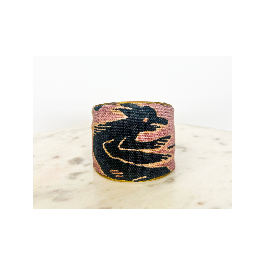 Studio product image of the Redd Dott “Zora” 2″ fabric cuff bracelet, showcasing sweeping black abstract swirls printed on textured dusty-rose cotton, punctuated by fine metallic-gold highlights and backed by a polished open brass interior, perched on a smooth marble surface.
