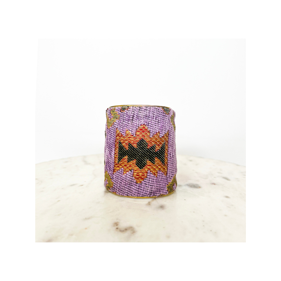 A handmade cuff bracelet, cylindrical and intricately beaded, features a striking geometric pattern in purple, orange, black, and green hues as it stands on a white surface.