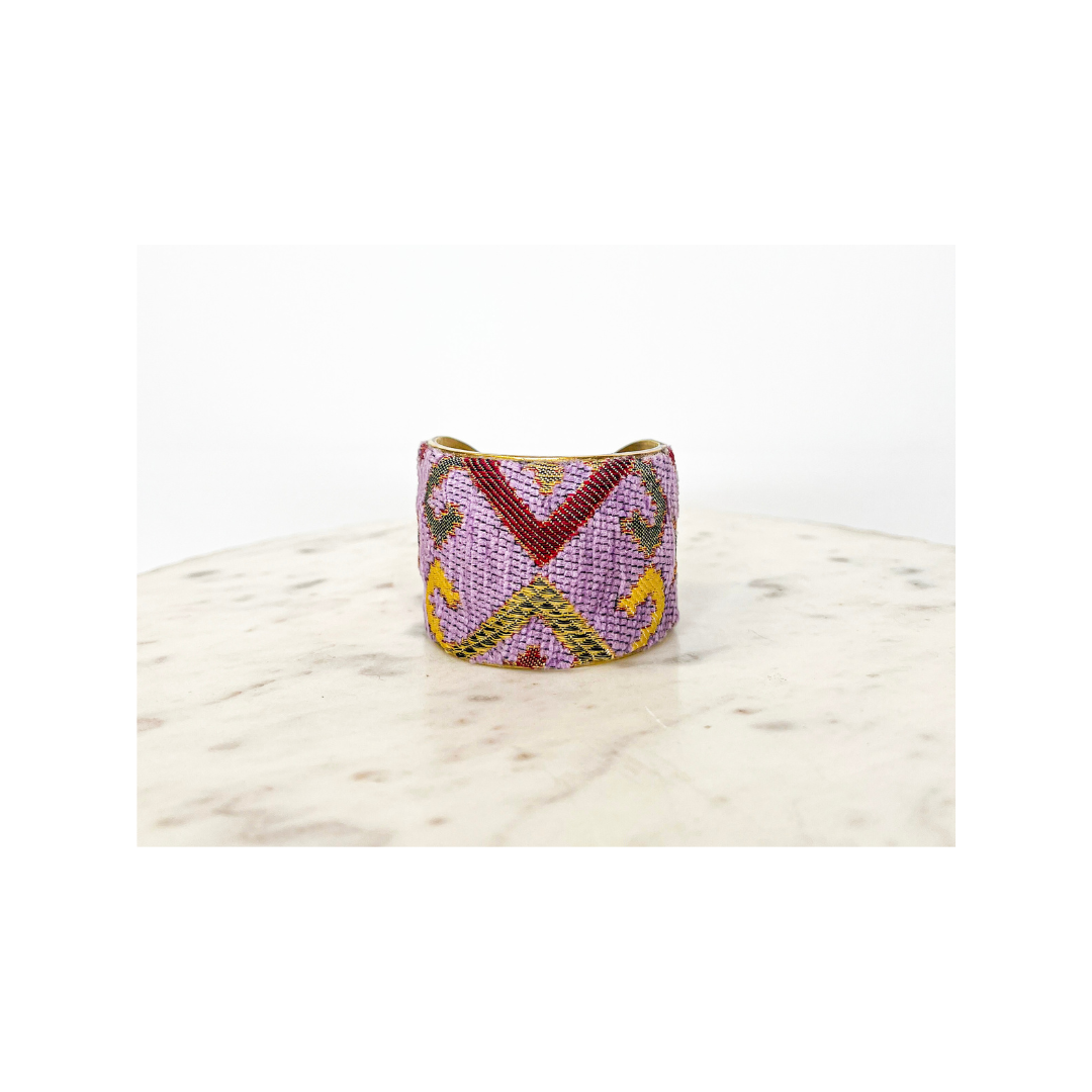 A handmade cuff bracelet with a pink, yellow, and red geometric pattern is displayed on a round, light-colored marble surface.