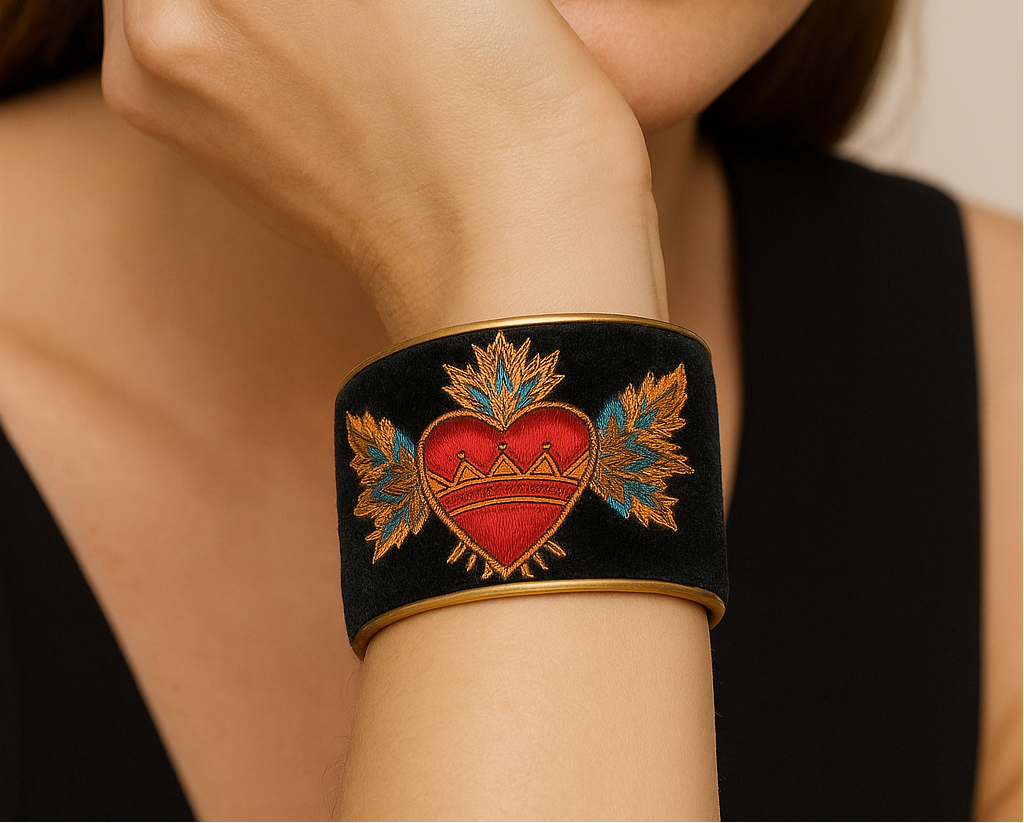 Close-up of a woman resting her face on her hand wearing the Redd Dott “Sacred Heart” 2″ cuff—black velvet bracelet with brass edging, featuring a bold embroidered red heart with a gold crown band, radiant gold rays, and teal-gold winged florals.