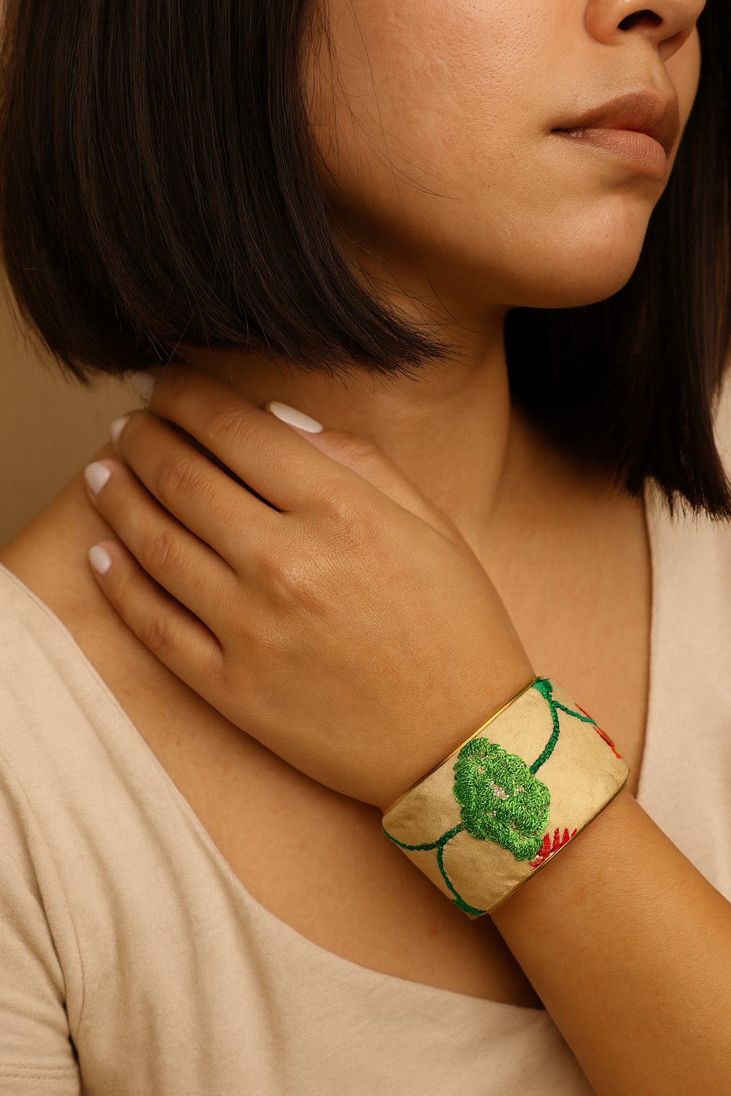 Close-up of a woman’s wrist and lower face, wearing the Redd Dott “Tia” 2″ wide fabric cuff. The pale champagne satin band is embroidered with a raised emerald-green rosette and winding teal vines with tiny coral accents, framed by a slim open-back brass shell as her hand rests at her collarbone.