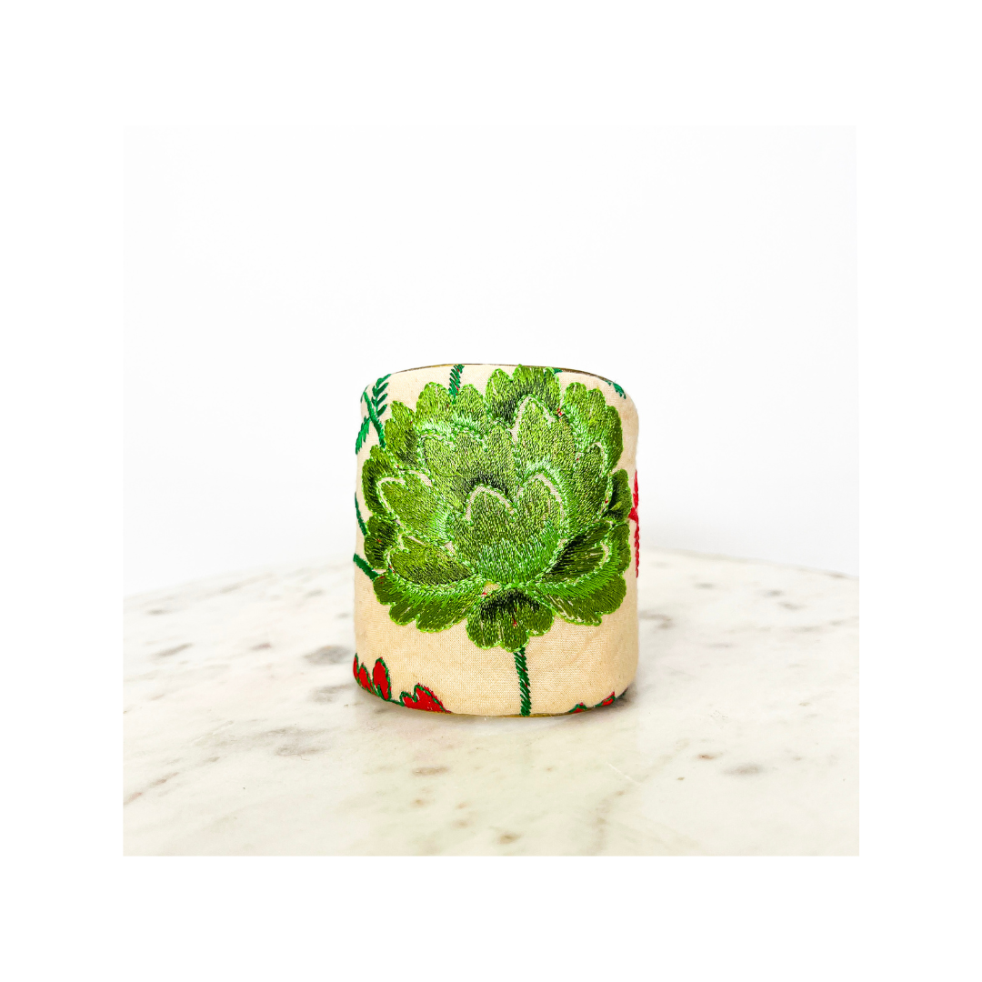 A beige cylindrical object with an embroidered green floral pattern, reminiscent of a floral cuff bracelet, sits on a marble surface against a plain white background.