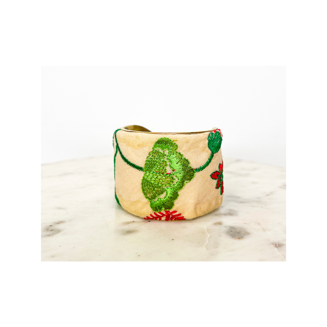A beige floral cuff bracelet with green and red embroidery, placed on a light marble surface.