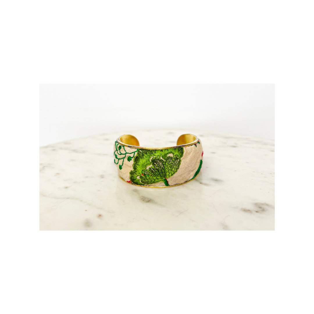 A wide, gold floral cuff bracelet with intricate green leaf designs on a light background, displayed on a marble surface.
