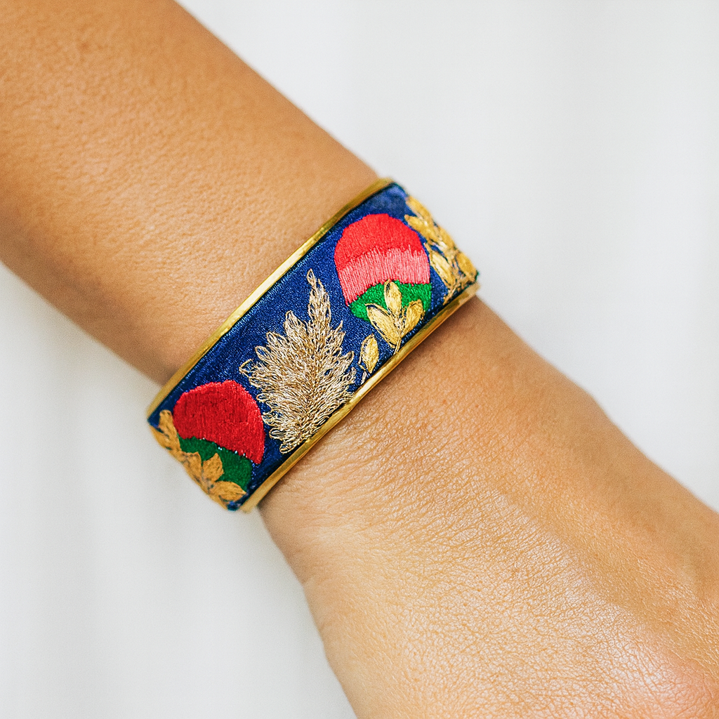 Close-up of a woman’s wrist wearing the Redd Dott “Trinika” 1″ fabric cuff. The royal-blue satin band is embroidered with bold red flower buds with green bases and metallic-gold wheat sprigs and leaves, framed by polished brass edges with an open back.