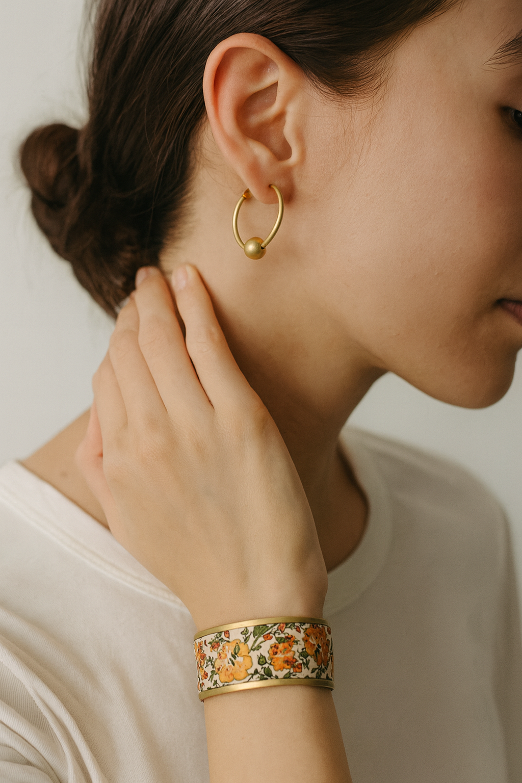 Close-up of a woman’s wrist and lower face, wearing the Redd Dott “Trixie” 1″ wide fabric cuff. The slim ivory band features a vintage-style floral print—buttercup yellow and apricot blossoms with leafy green vines—framed by a slender open-back brass shell as her hand touches her ear.