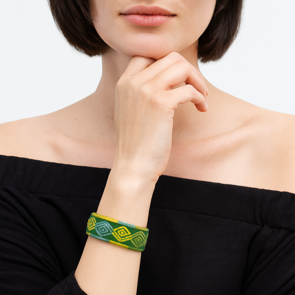 Close-up of a woman’s wrist and lower face, wearing the Redd Dott “Tz’unun” 1″ wide fabric cuff. The slim band features tightly stitched geometric diamond motifs in ombré greens—jade to forest—with pops of citrus yellow along the border, set within a slender open-back brass frame as her hand rests beneath her chin.