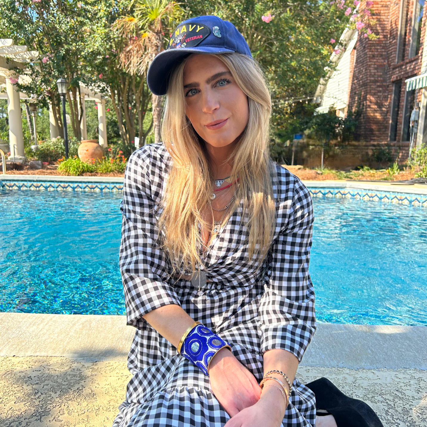 A person sits by the pool, wearing a checkered dress, a navy cap, and blue and gold jewelry, including a Spring Floral Cuff Bracelet. In the background, you can see a brick building and lush greenery.