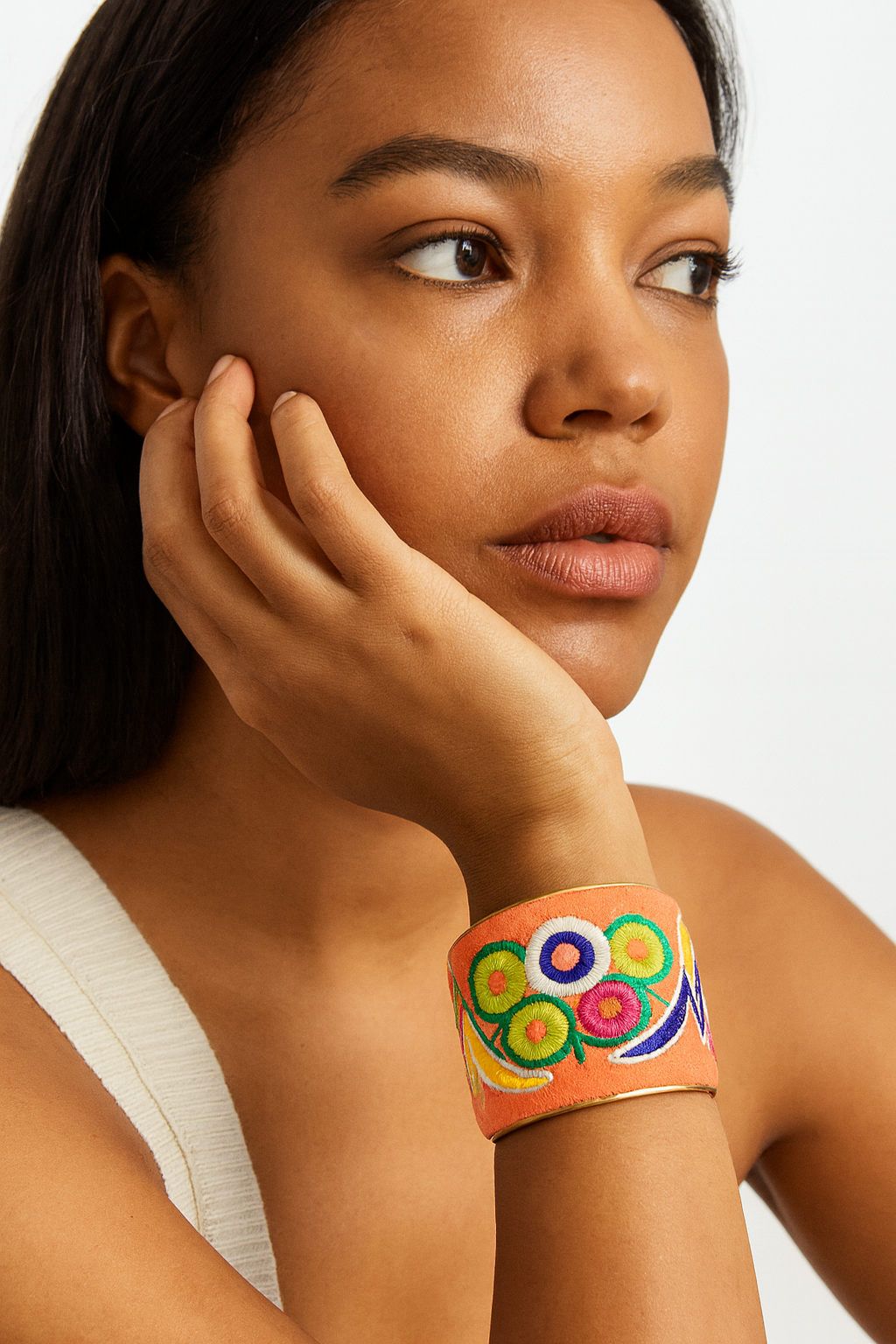 Close-up of a woman’s wrist and lower face, wearing the Redd Dott “Vanna” 2″ wide fabric cuff. The sunlit tangerine cotton base is embroidered with concentric circle floral motifs in emerald green, white, fuchsia, and lemon yellow, accented by swirling cobalt and gold stitch lines, all set within a slender open-back brass frame as her hand gently supports her cheek.