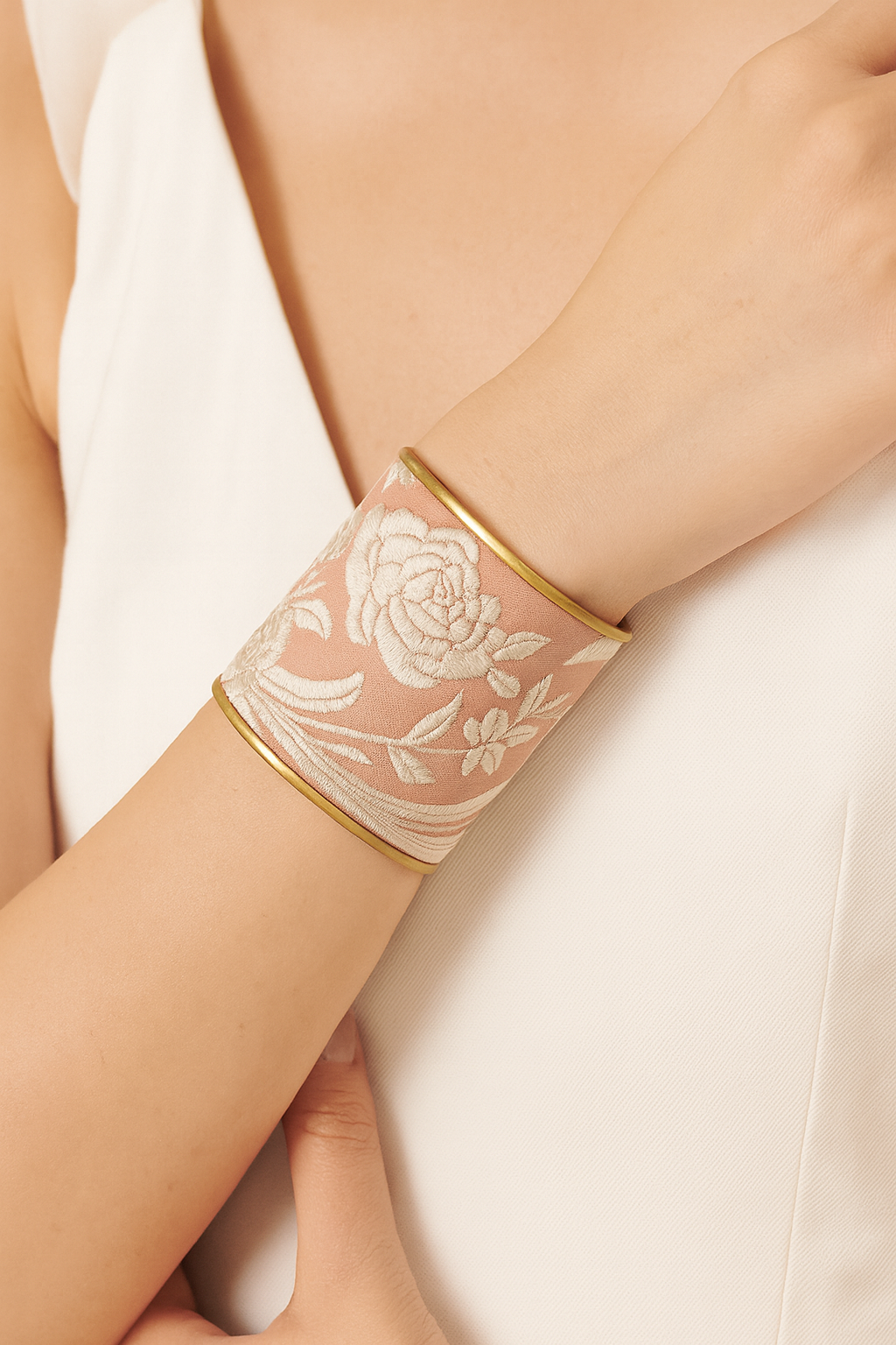 Close-up of a woman’s wrist and lower face, wearing the Redd Dott “Zaria” 3″ wide fabric cuff. The soft dusty-rose cotton is richly embroidered with raised ivory floral blooms and trailing vines, set within smooth matte-brass edges as her hand gently rests against her chest.