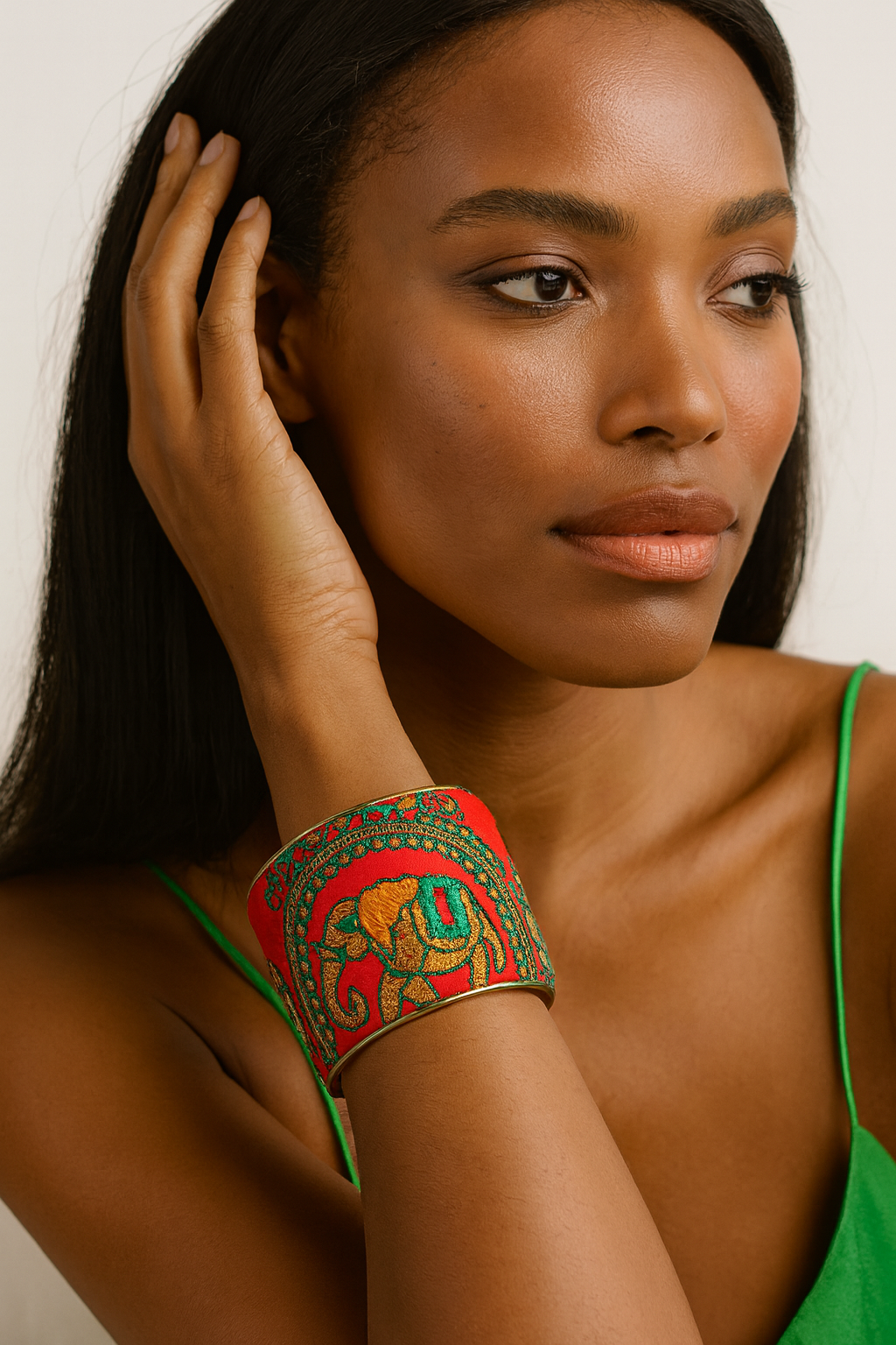 Close-up of a woman’s wrist and lower face, wearing the Redd Dott “Zelda” 3″ wide fabric cuff. The bold scarlet cotton base is richly embroidered with a metallic-gold elephant silhouette framed by twisting emerald-green vines and red stitch-dots, all set in a gently curved open-back brass shell as her fingers brush back her hair.
