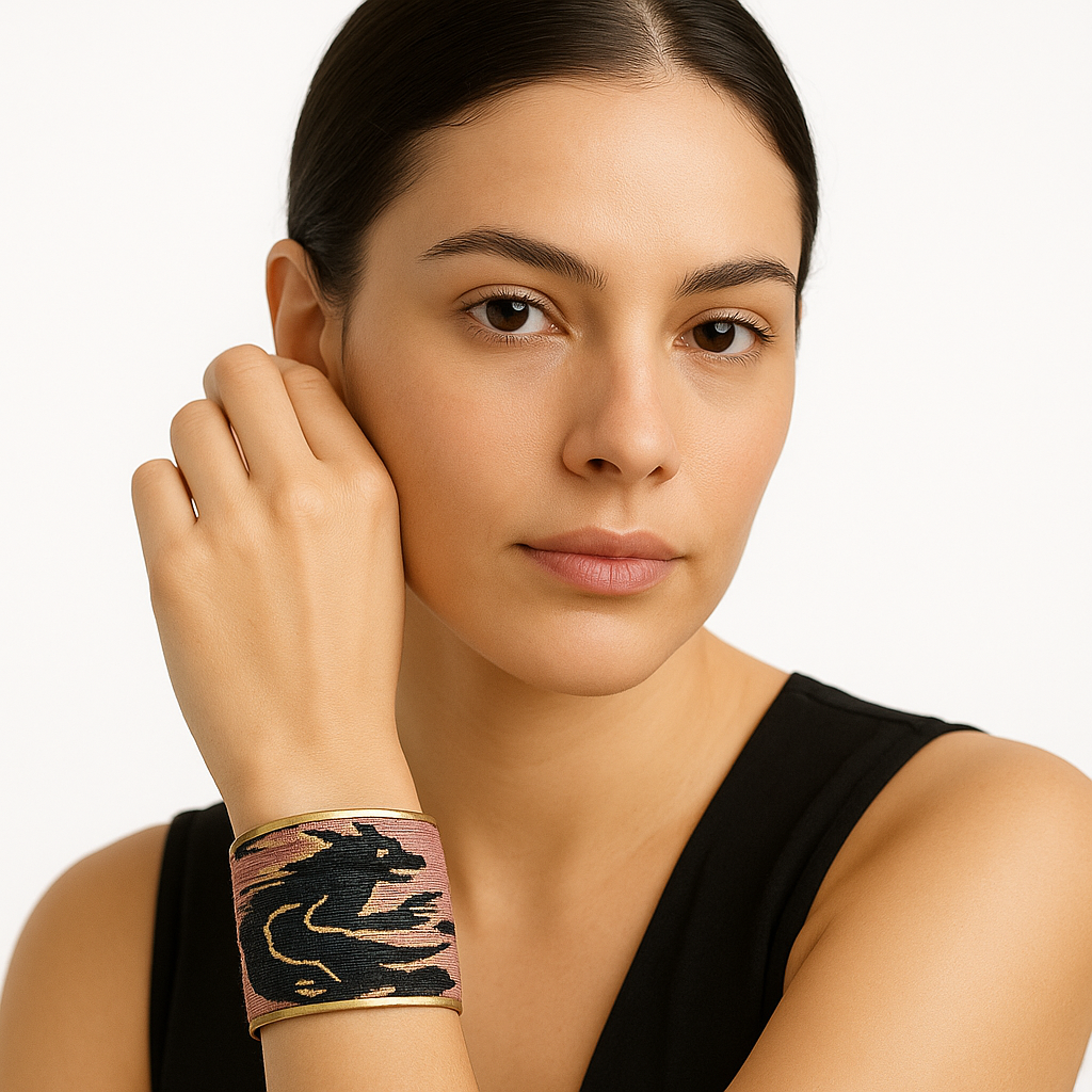 Close-up of a woman’s lower face and wrist, wearing the Redd Dott “Zora” 3″ wide fabric cuff. The cuff’s dusty-rose cotton weave is patterned with bold, inky black brushstroke swirls and interspersed with subtle metallic-gold streaks, all framed by an open-back brass shell as her hand gently rests against her temple.