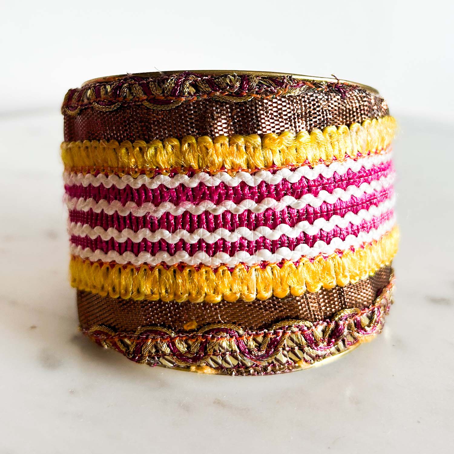 A standalone 2-inch wide raw brass cuff bracelet wrapped in bronze metallic fabric, featuring embroidered fuchsia and white stripes, bold yellow looped stitching, and braided metallic trim on the top and bottom edges, shown against a white background.