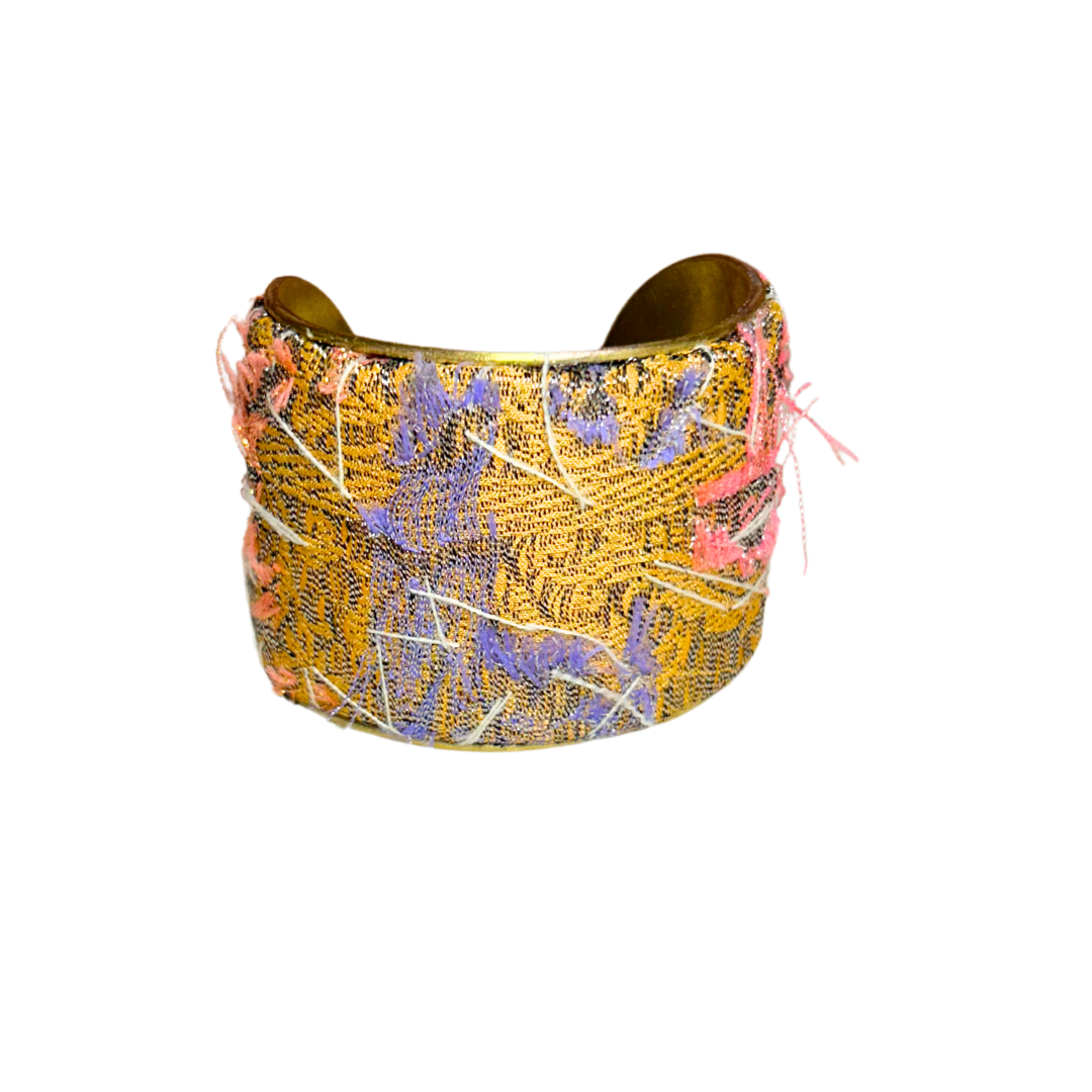 A handmade brass cuff bracelet; crafted and stamped in Shreveport, LA. Wrapped in golden yellow fabric with loose coral, purple, ivory, and dark threads, giving it a textured, distressed look perfect for a rustic cuff bracelet style.