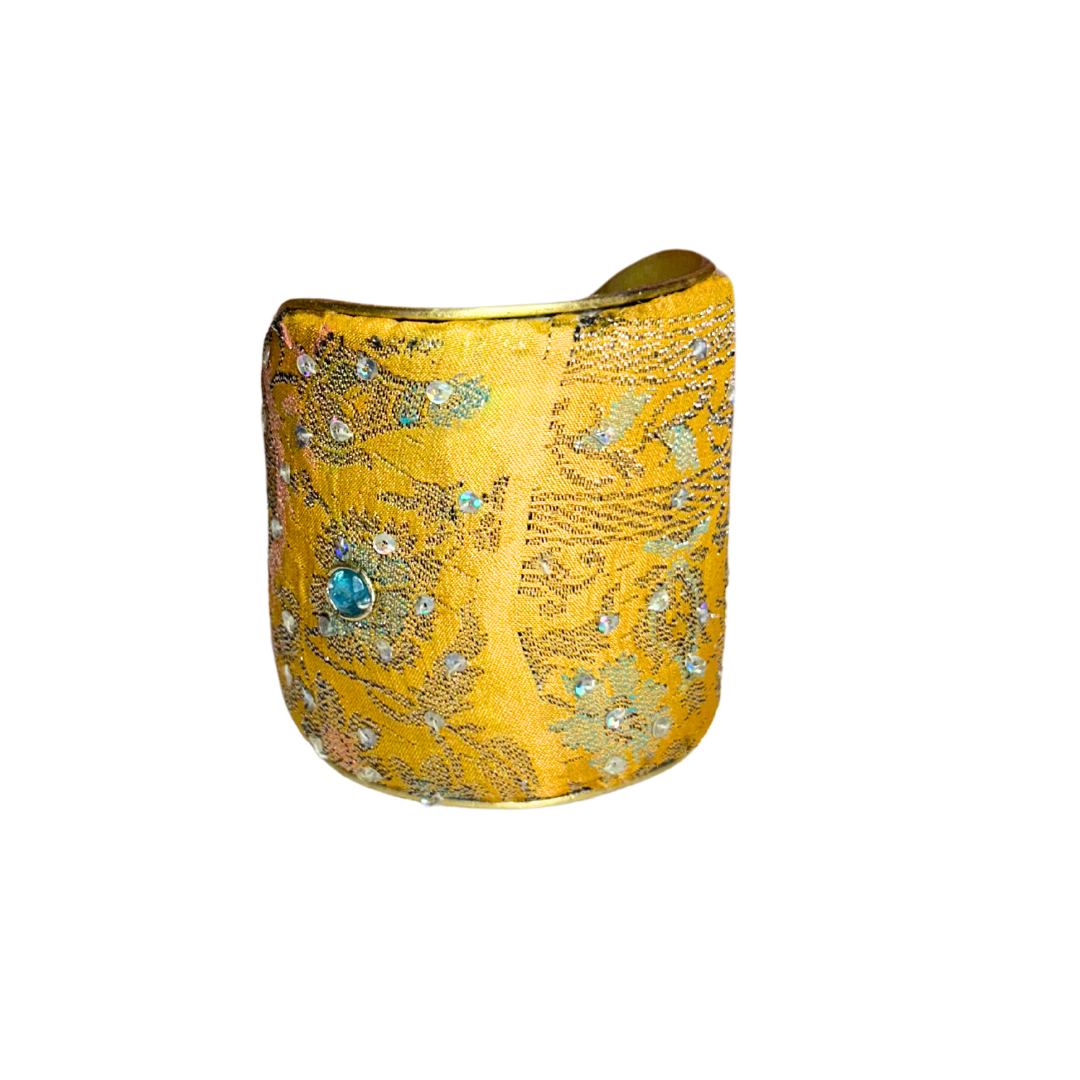 A handmade brass cuff bracelet; crafted and stamped in Shreveport, LA. Wrapped in golden yellow fabric with loose coral, purple, ivory, and dark threads, giving it a textured, distressed look perfect for a rustic cuff bracelet style.