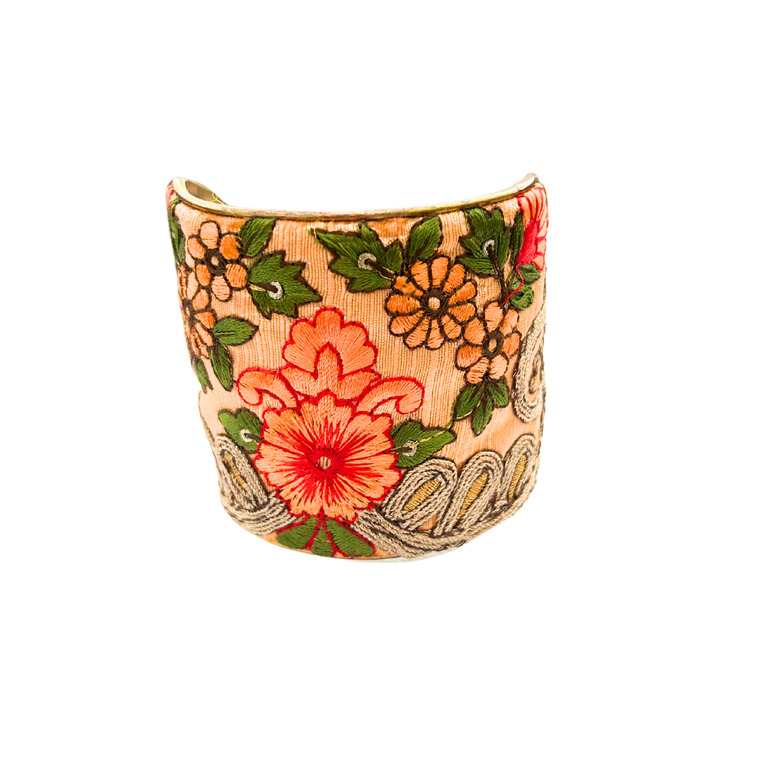 A handcrafted brass cuff bracelet by Redd Dott Jewelry featuring embroidered coral and gold flowers with green leaves on a peach background. A bold and elegant luxury flower bracelet design.