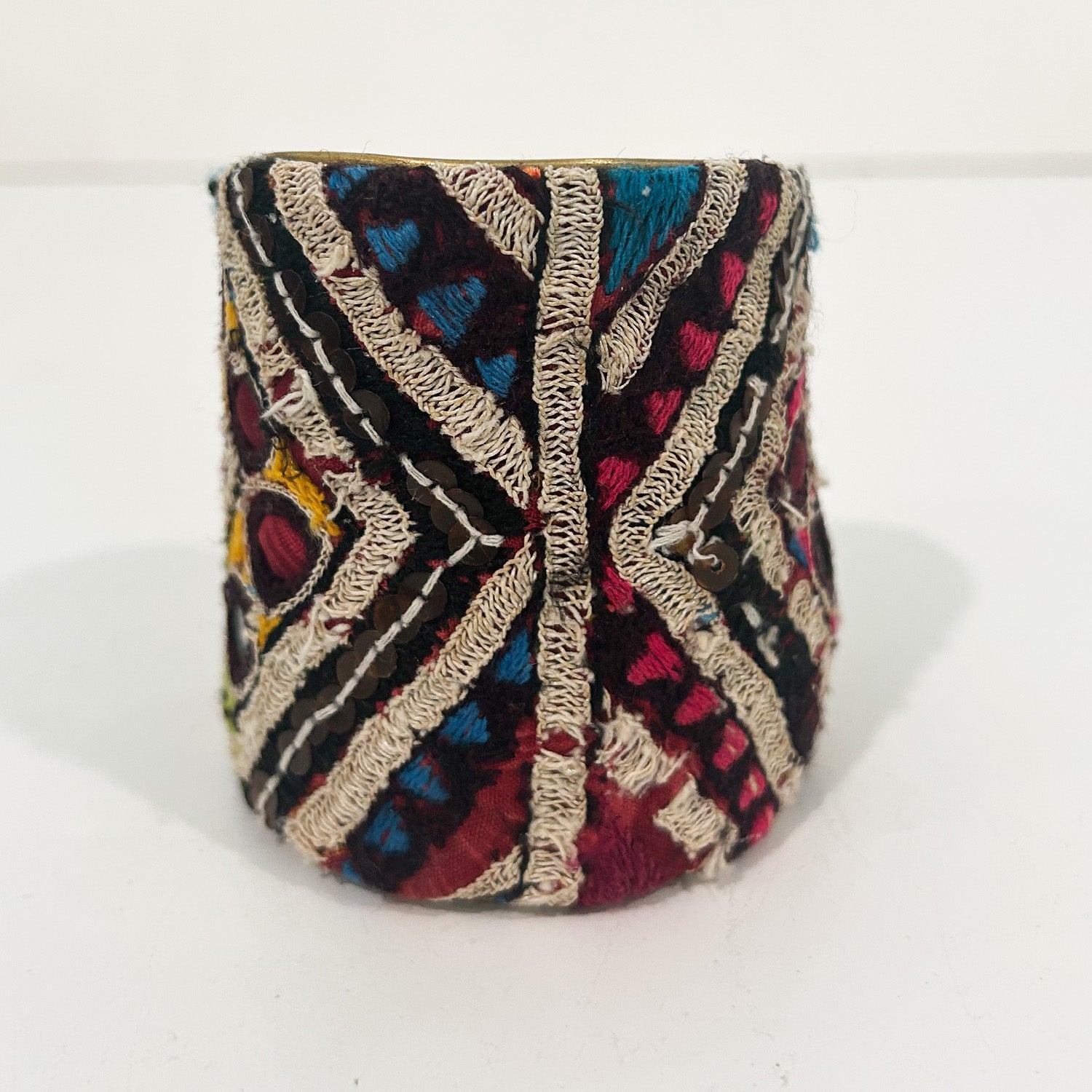 A 3-inch wide raw brass cuff bracelet wrapped in hand-embroidered velvet fabric displaying bold diamond and chevron motifs in crimson, teal, mustard, and cream threads with scattered sequin embellishments, set against a clean white background.