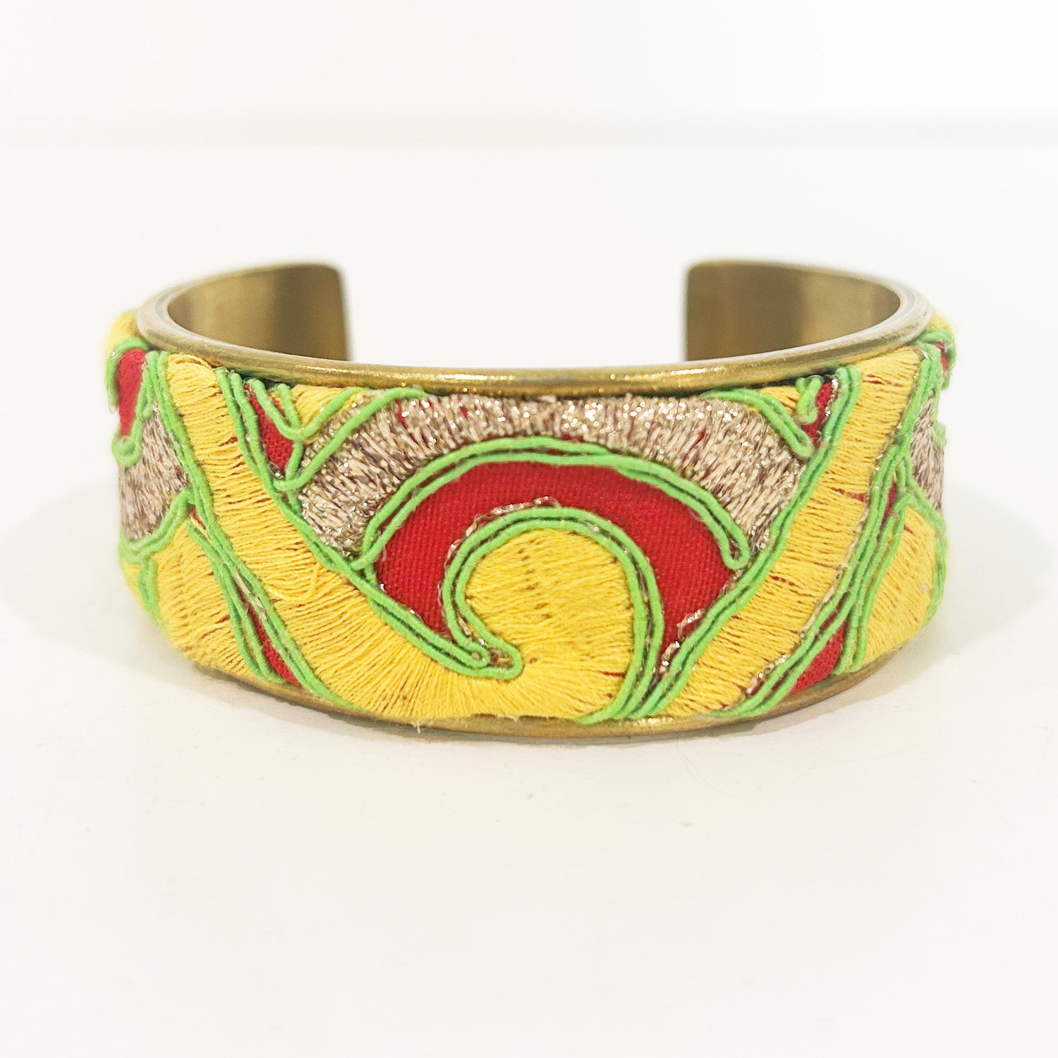 A standalone 1-inch wide raw brass cuff bracelet wrapped in red linen fabric, featuring hand-embroidered swirling velvet motifs in bright yellow, metallic gold thread accents, and neon green outlines, photographed against a white background.