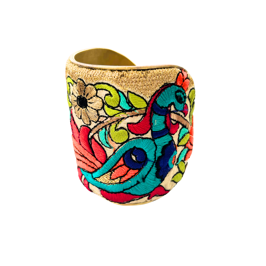 A handcrafted brass cuff bracelet by Redd Dott Jewelry with an embroidered peacock and floral motif in vibrant teal, coral, and gold. A bold, colorful statement cuff bracelet with a golden fabric base and vivid threadwork.
