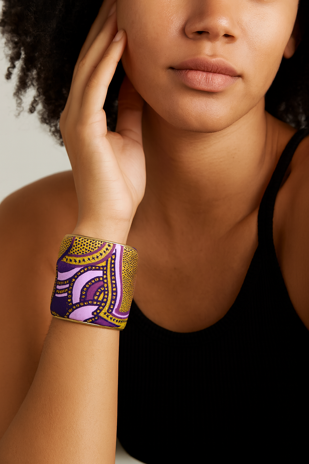 Close-up of a woman’s wrist and lower face, wearing the Redd Dott “Life of the Mardi” 3″ wide fabric cuff. The cuff shows undulating lavender and deep purple arcs veined with clusters of tiny metallic-gold dots atop soft, textured cotton, its open-back brass frame gently cradling her wrist.