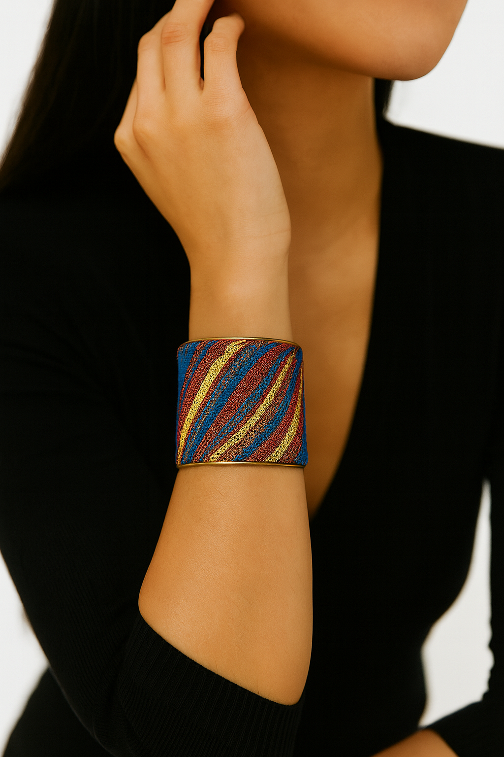Close-up portrait of a female model wearing a 3-inch wide blue, red, and gold striped brass cuff bracelet; the open-back ends peek out as she rests her hand against her face.