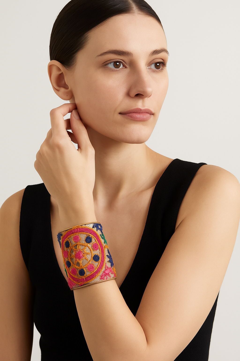 A woman in a black sleeveless top touches her ear, showcasing a 3-inch wide brass cuff bracelet wrapped in linen with hand-embroidered medallion motifs in fuchsia, orange, green, and blue, with a smooth brass interior visible at the edges.