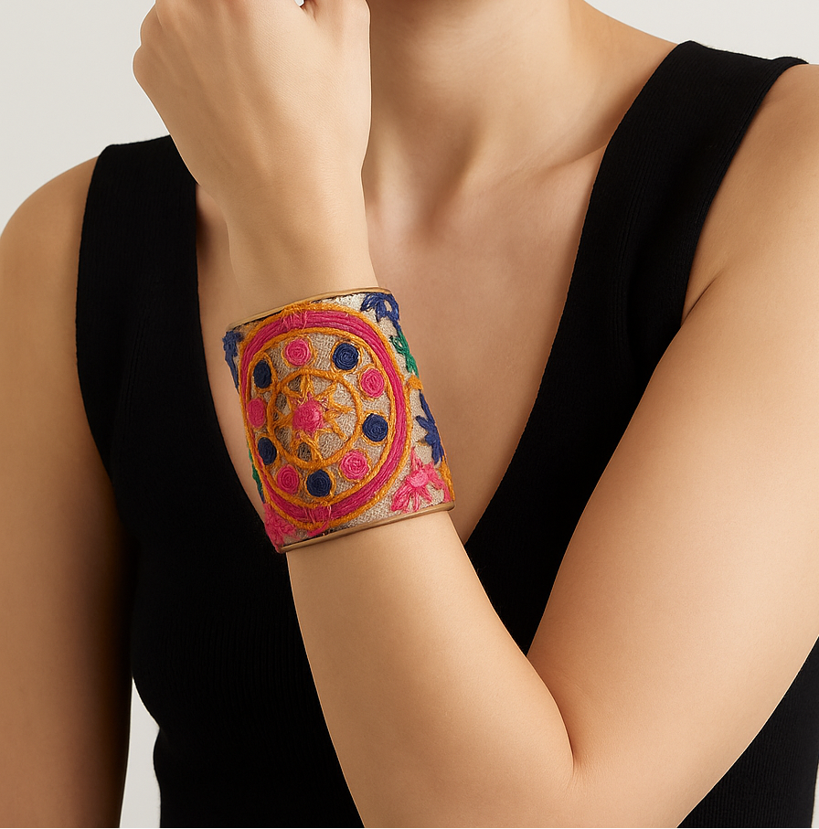 A woman in a black sleeveless top touches her ear, showcasing a 3-inch wide brass cuff bracelet wrapped in linen with hand-embroidered medallion motifs in fuchsia, orange, green, and blue, with a smooth brass interior visible at the edges.