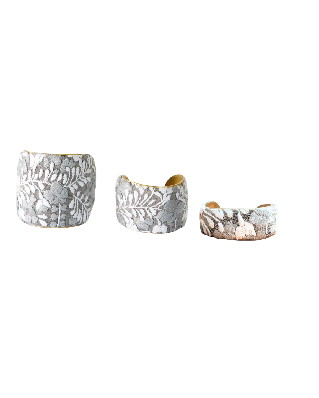 Trio of Niebla brass cuff bracelets in 1”, 2”, and 3” widths featuring mist-gray and white leaf embroidery on brass bases, arranged side by side against a white background.