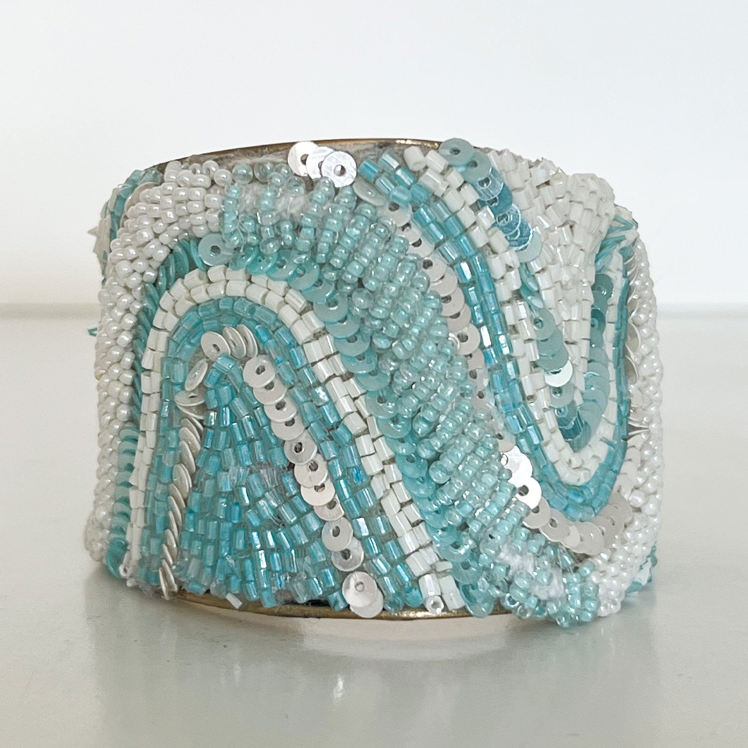 A 2-inch brass cuff bracelet from Redd Dott Jewelry covered in hand-stitched aqua and pearl white seed beads with layered sequins forming flowing wave patterns—an eye-catching statement cuff bracelet and piece of inspirational jewelry.
