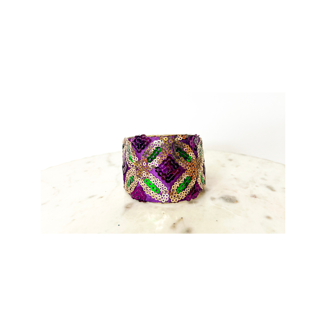 A studio image of a one of a kind jewelry brass cuff bracelet for women—a 2-inch wide, open-back design wrapped in purple fabric with gold and green sequin embroidery in diamond motifs, shown on a white marble surface.