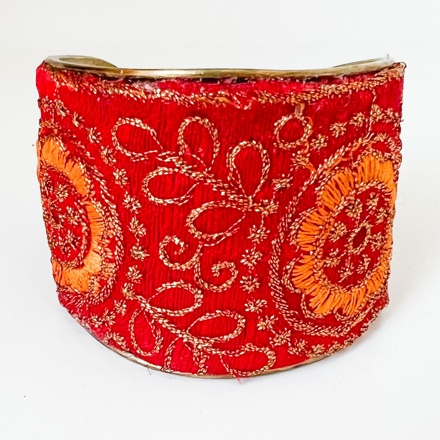 A studio image of a one of a kind jewelry brass cuff bracelet for women—a 2-inch wide, open-back brass cuff wrapped in hand-embroidered red fabric with orange and gold blossom embroidery, displayed on a white marble surface.