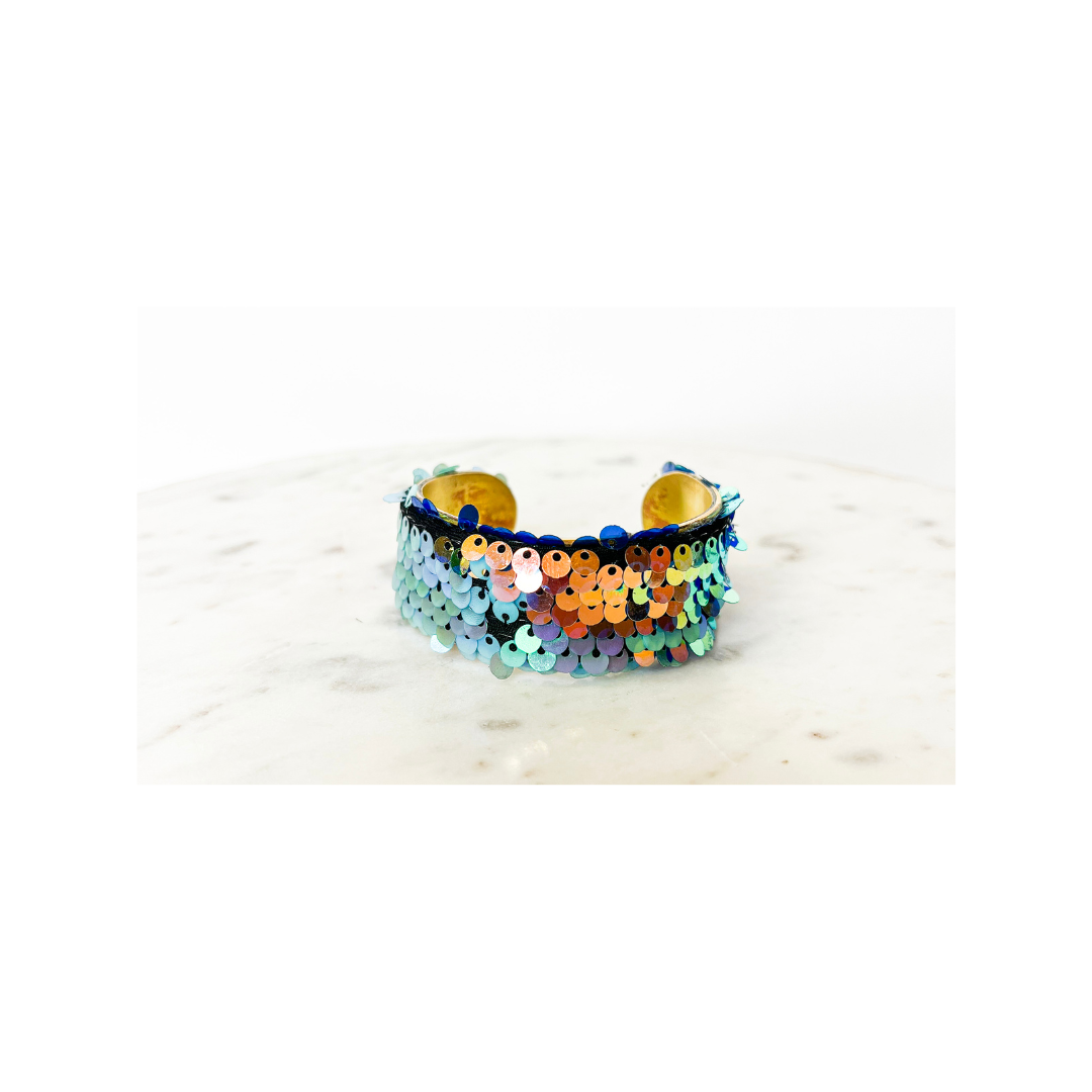 A studio image of a one of a kind jewelry brass cuff bracelet for women—a 1-inch wide open-back design wrapped in gradient iridescent sequins over a black fabric base, displayed on a white marble surface.
