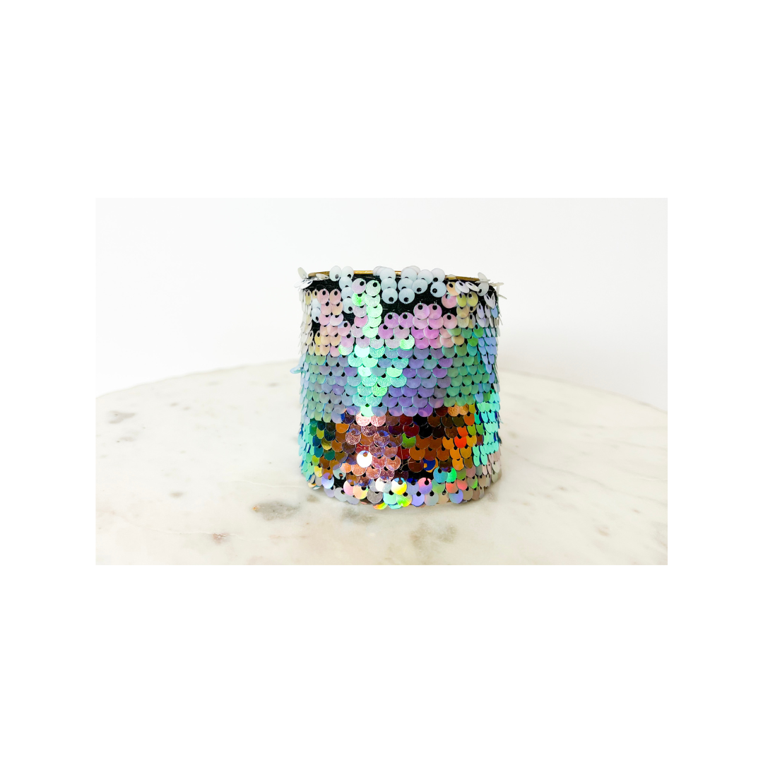 A studio image of a one of a kind jewelry brass cuff bracelet for women—a 3-inch wide open-back design wrapped in shimmering sequins that shift from pink to green to orange, showcased on a white marble surface.