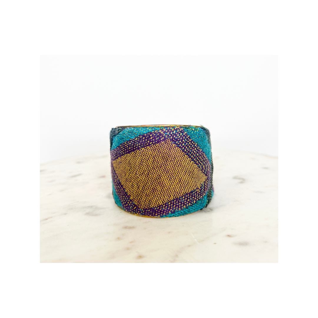 A studio image of a one of a kind jewelry brass cuff bracelet for women—a 2-inch wide open-back design wrapped in teal, amethyst, and gold woven fabric featuring a diamond pattern, displayed on a white marble surface.