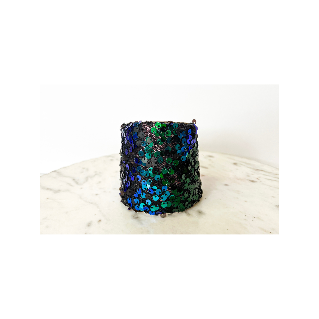 A studio image of a one of a kind jewelry brass cuff bracelet for women—a 3-inch wide, open-back design wrapped in black fabric densely covered with green and blue iridescent sequins, displayed on a white marble surface.