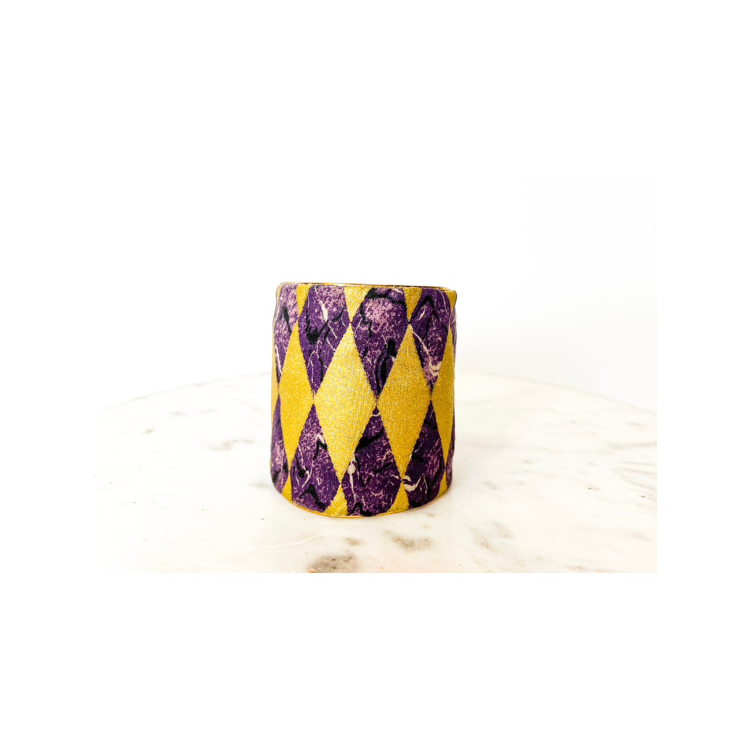 A studio image of a one of a kind jewelry brass cuff bracelet for women—a 3-inch wide, open-back design wrapped in jacquard fabric with metallic gold and purple diamond patterns, displayed on a white marble surface.