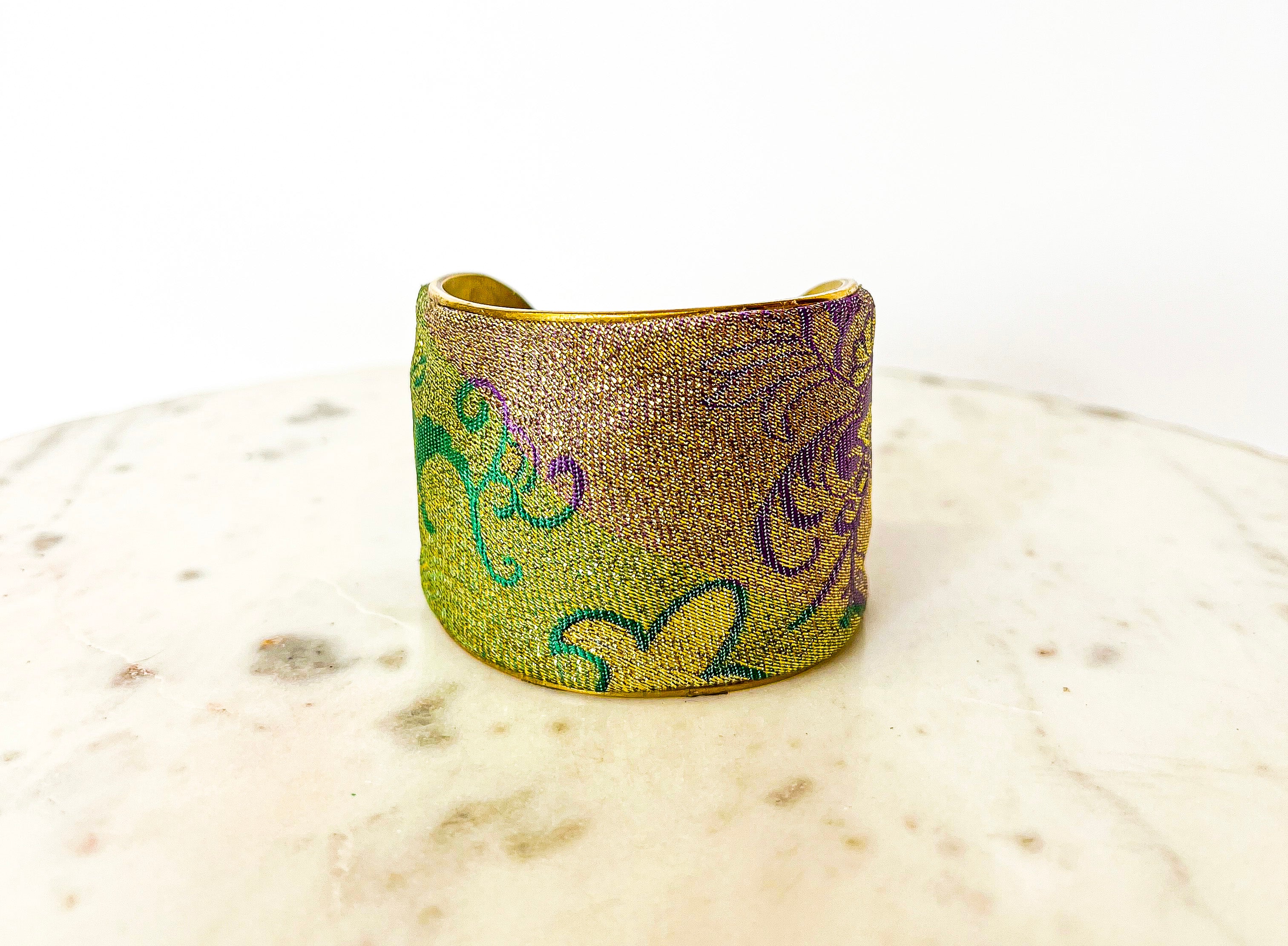 A studio image of a brass cuff bracelet for women—available in 1-inch, 2-inch, and 3-inch widths—wrapped in metallic gold, emerald green, and purple tapestry fabric with swirling carnival patterns, displayed on a white marble surface.