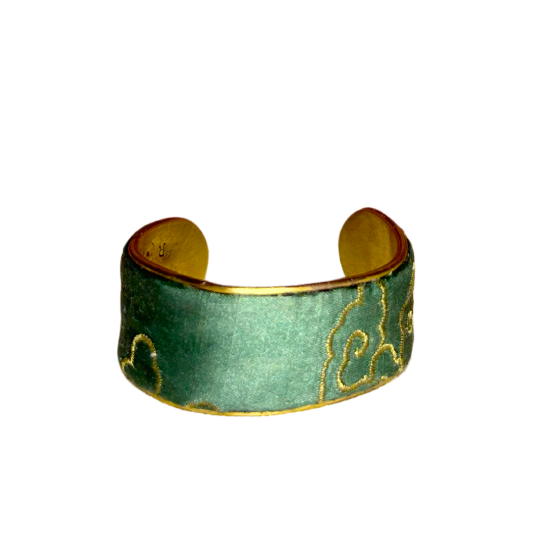 The Tillie brass cuff bracelet features intricate embroidered symbols in sky blue and gold on a sage green satin base. This bold power bracelet merges the elegance of inspirational jewelry with timeless brass craftsmanship.