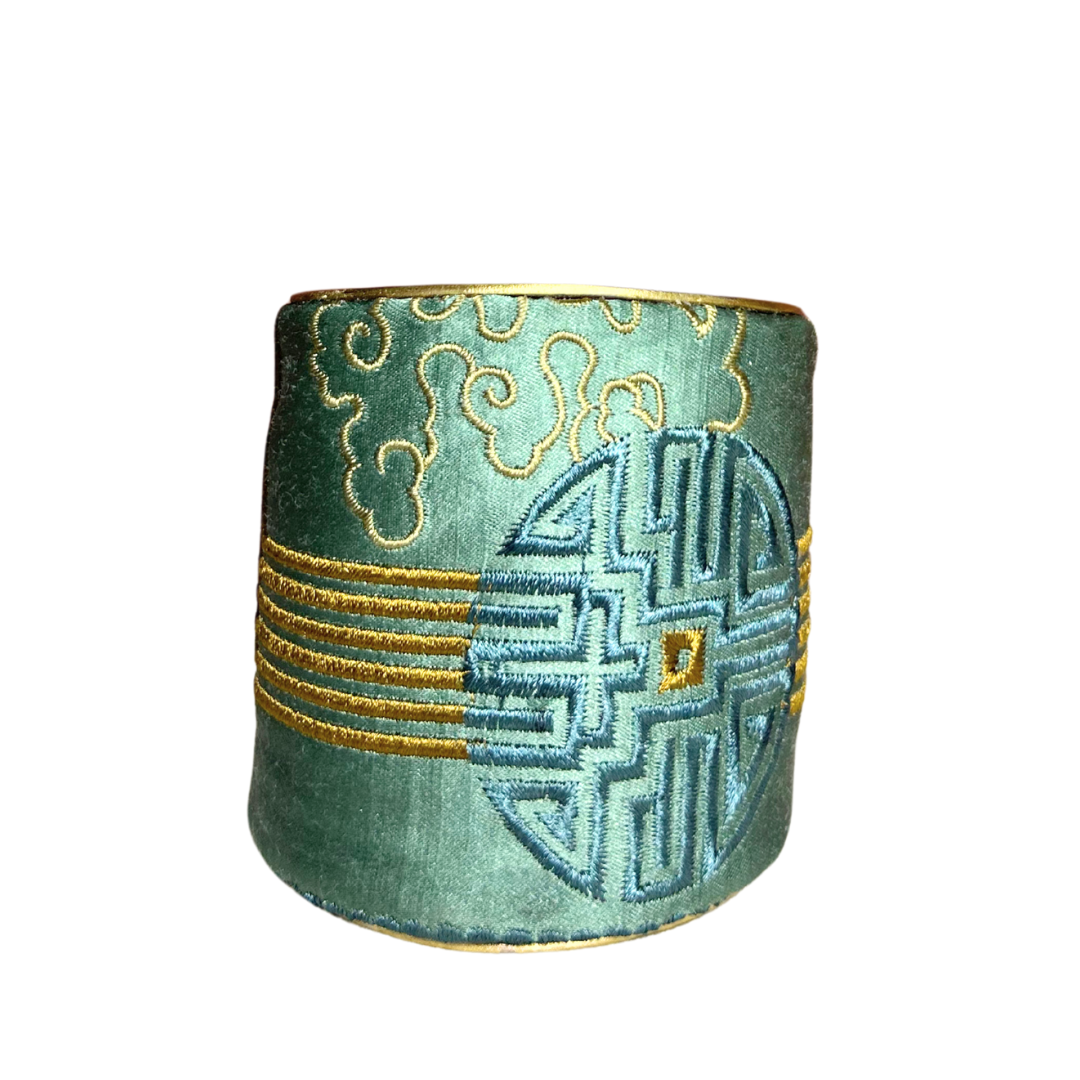 The Tillie brass cuff bracelet features intricate embroidered symbols in sky blue and gold on a sage green satin base. This bold power bracelet merges the elegance of inspirational jewelry with timeless brass craftsmanship.