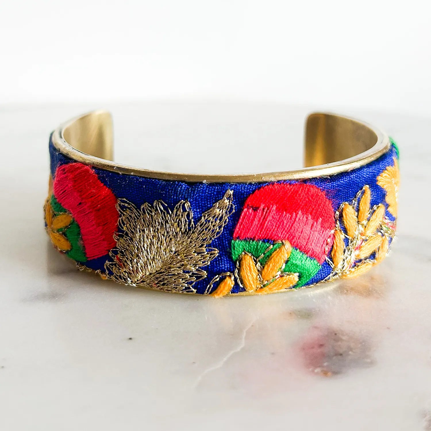 A 1-inch brass cuff bracelet featuring a vibrant blue fabric embroidered with red florals, green leaves, and golden threads—an elegant piece of embroidered jewelry and inspirational jewelry handcrafted by Redd Dott Jewelry.